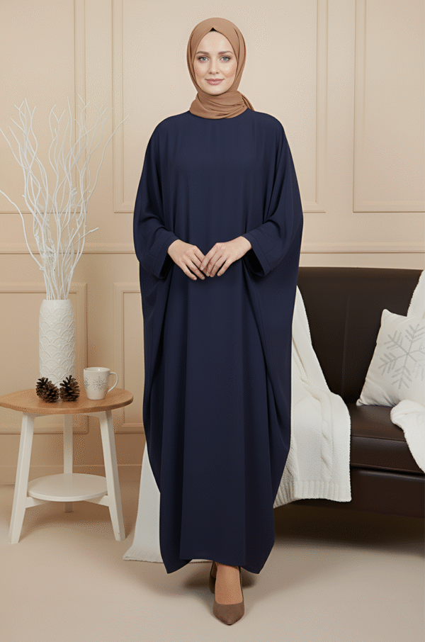 Beautiful woman wearing navy blue abaya and beige hijab, standing indoors with cozy decor.