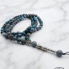 Blue and gray beaded mala necklace with silver pendant on marble surface.