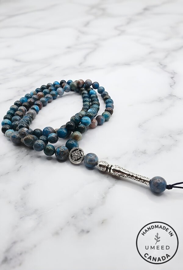 Blue and gray beaded mala necklace with silver pendant on marble surface.