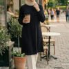 Elegant woman in modest black dress and beige hijab holding coffee outside a cafe.