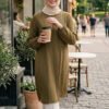 A smiling woman in a beige hijab and olive green dress holding coffee outside a café in a lively cit.