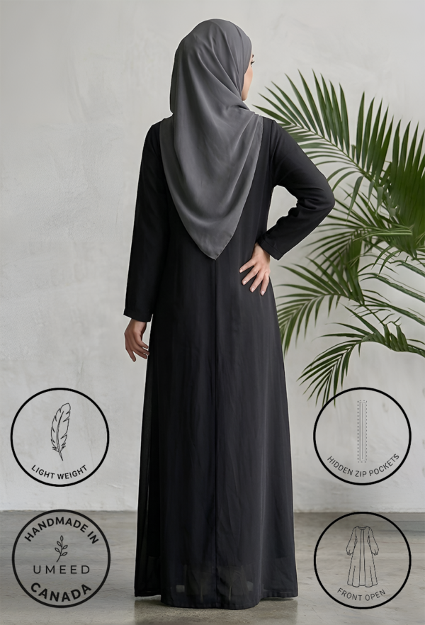 Elegant woman in black abaya with hijab standing near a green plant.